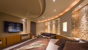 A modern hotel room with a large bed, a flat-screen TV mounted on the wall, a seating area with a sofa and armchairs, and a decorative wall panel. - Hotel C. Chiba Shiroi (Shiroi)
