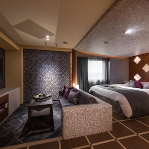 A hotel room with a bed, a sofa, a small table with a tray of fruits, and a patterned wall. - Hotel C. Chiba Shiroi (Shiroi)