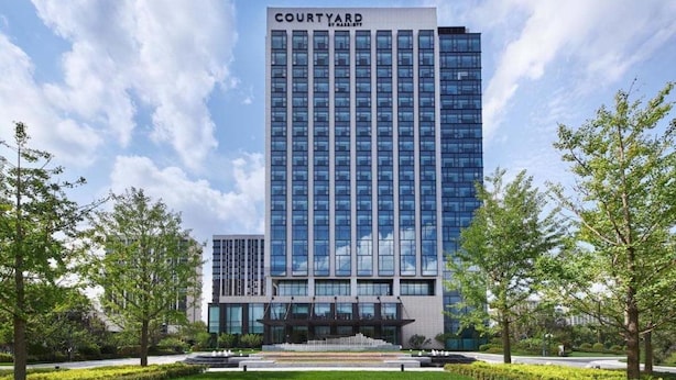 En moderne hotelbygning med et tydeligt 'Courtyard' skilt, omgivet af velplejede grønne områder og en klar himmel.