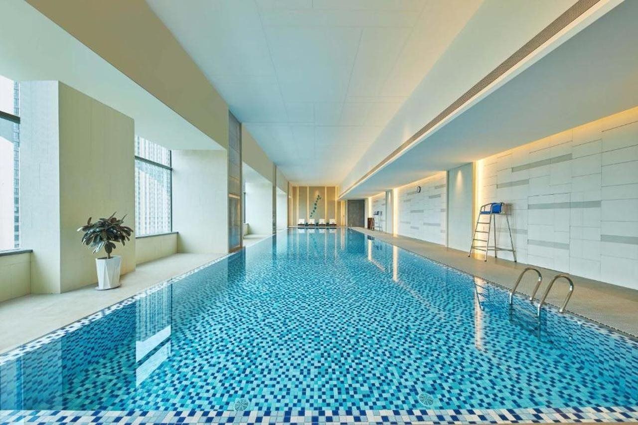 A long indoor swimming pool with clear blue water, a potted plant, and a ladder on one side.