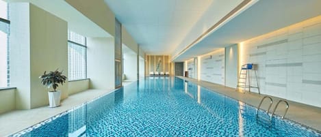 A long indoor swimming pool with clear blue water, a potted plant, and a ladder on one side.