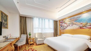 Habitación de hotel con una cama, un escritorio, una silla, una mesita con una planta y un cuadro en la pared.