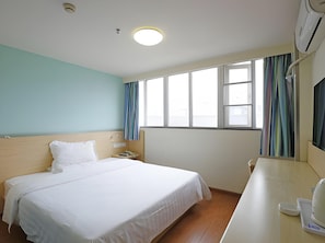 Hotellihuone, jossa on sänky, yöpöytä, lamppu, ikkuna verhoilla ja ilmastointilaite.