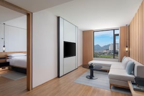 Quarto de hotel moderno com cama, sofá, TV fixada na parede e uma ampla janela com vista para a cidade.