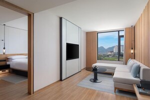 Quarto de hotel moderno com cama, sofá, TV fixada na parede e uma ampla janela com vista para a cidade.