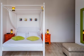 A four-poster bed with white linens and two pillows, flanked by two nightstands with lamps, in a room with a wooden floor and a green-framed mirror. - Eiriana Luxury Suites (Milos)