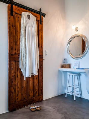 A room with a wooden sliding door, a white dress hanging on it, a round mirror, a small stool, and a pair of sandals on the floor. - Eiriana Luxury Suites (Milos)