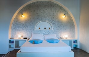 Une chambre à coucher avec un lit blanc, deux oreillers bleus, une tête de lit blanche et deux tables de chevet avec des lampes.