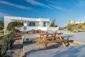 Un bâtiment blanc avec des volets bleus, une table de pique-nique et, au fond, une tour en pierre.