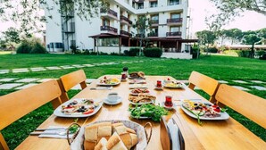 An outdoor dining area with a table set for a meal, surrounded by chairs, with a building in the background. - Troya Otel (Ezine)