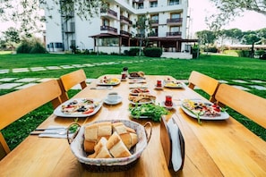 An outdoor dining area with a table set for a meal, surrounded by chairs, with a building in the background. - Troya Otel (Ezine)