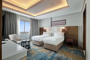 Ein Hotelzimmer mit zwei Betten, einem Schreibtisch, einem Sessel, einem kleinen Tisch und einem großen Fenster.