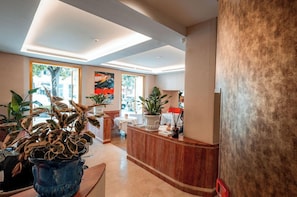 A hotel lobby with a reception desk, potted plants, and large windows allowing natural light. - Hotel Via Veneto (Rome)