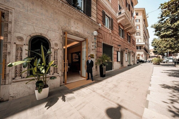 A hotel entrance with a man standing in the doorway, a potted plant, and a clear sky. - Hotel Via Veneto (Rome)