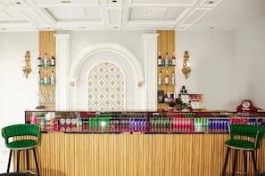 Una zona de bar con una barra de madera, dos sillones tapizados de verde y estantes con botellas y latas.
