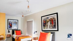 Quarto de hotel com duas camas, uma escrivaninha, uma cadeira, um guarda-roupa e obras de arte nas paredes.