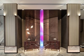 Un hall con suelo de mármol, una columna central con un elemento decorativo de luz vertical y dos mostradores de recepción con lámparas.