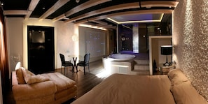 Una habitación de hotel moderna con una cama grande, una zona de estar con dos sillones, una mesita y una bañera, además de una pared con un diseño texturizado.