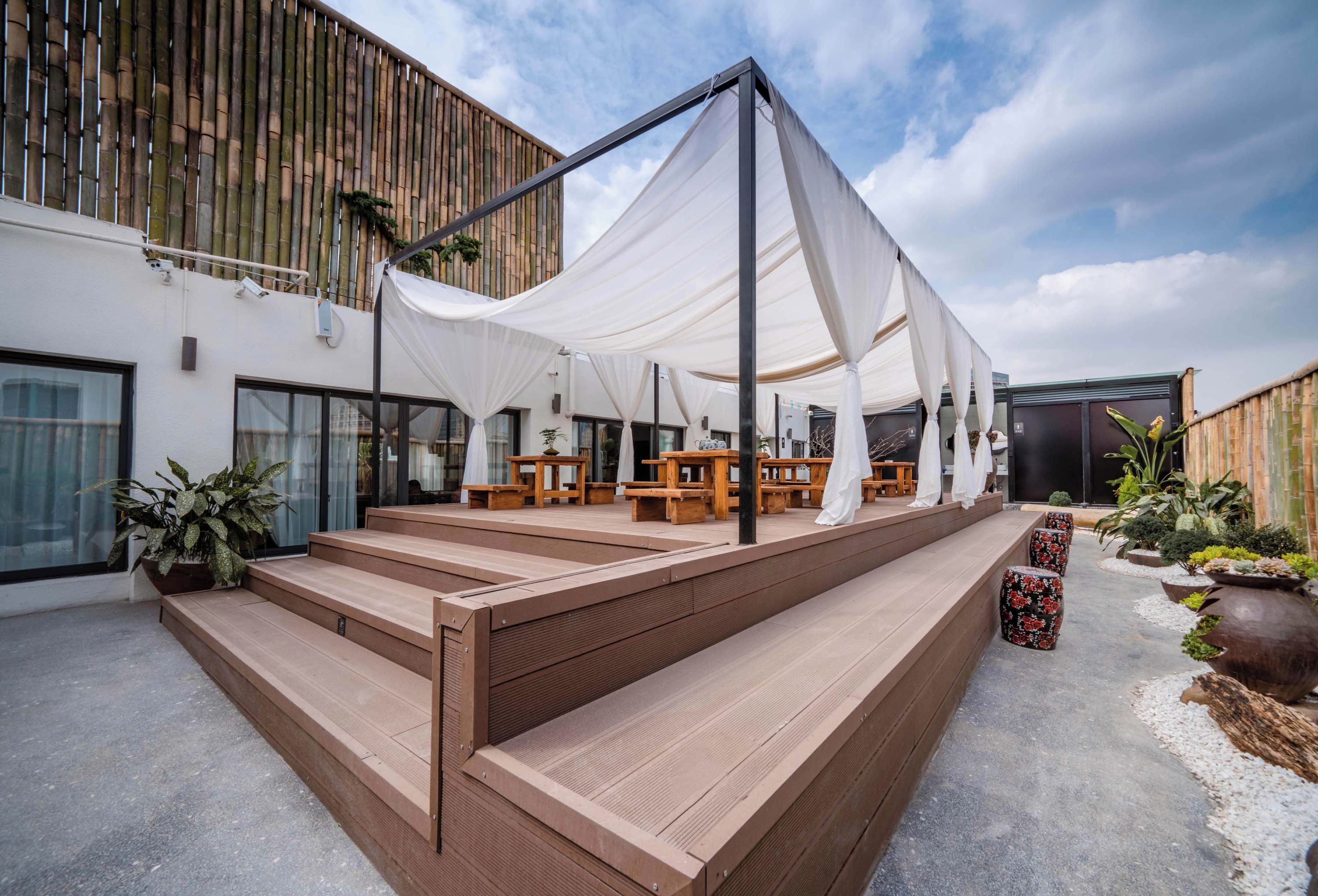 Un área de estar al aire libre con bancos y mesas de madera, un toldo blanco y una pared de bambú.