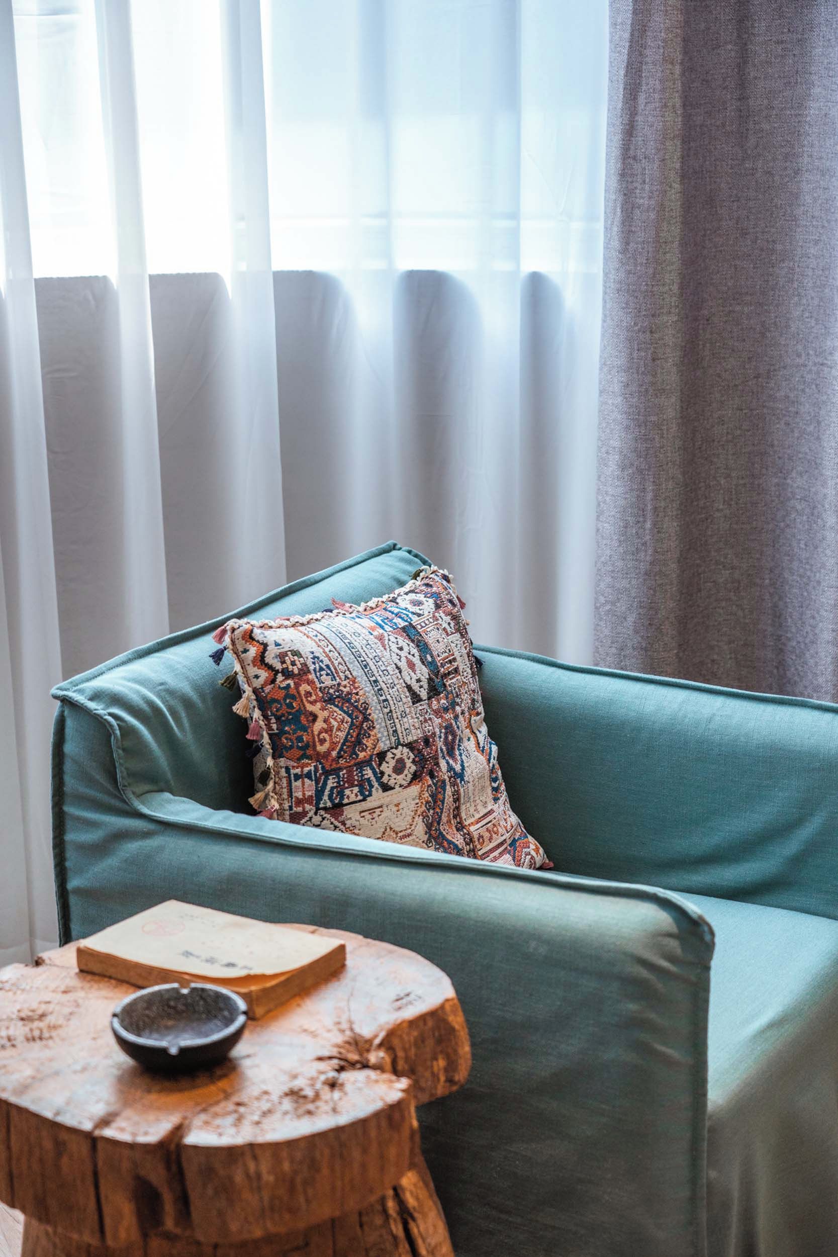 Um sofá verde-água com uma almofada estampada, uma mesinha de madeira com um livro e uma tigela, e cortinas leves.