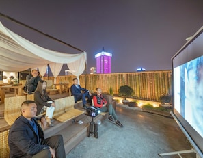 A group of people seated on outdoor furniture in a covered area with a cityscape view, including a brightly lit tower.