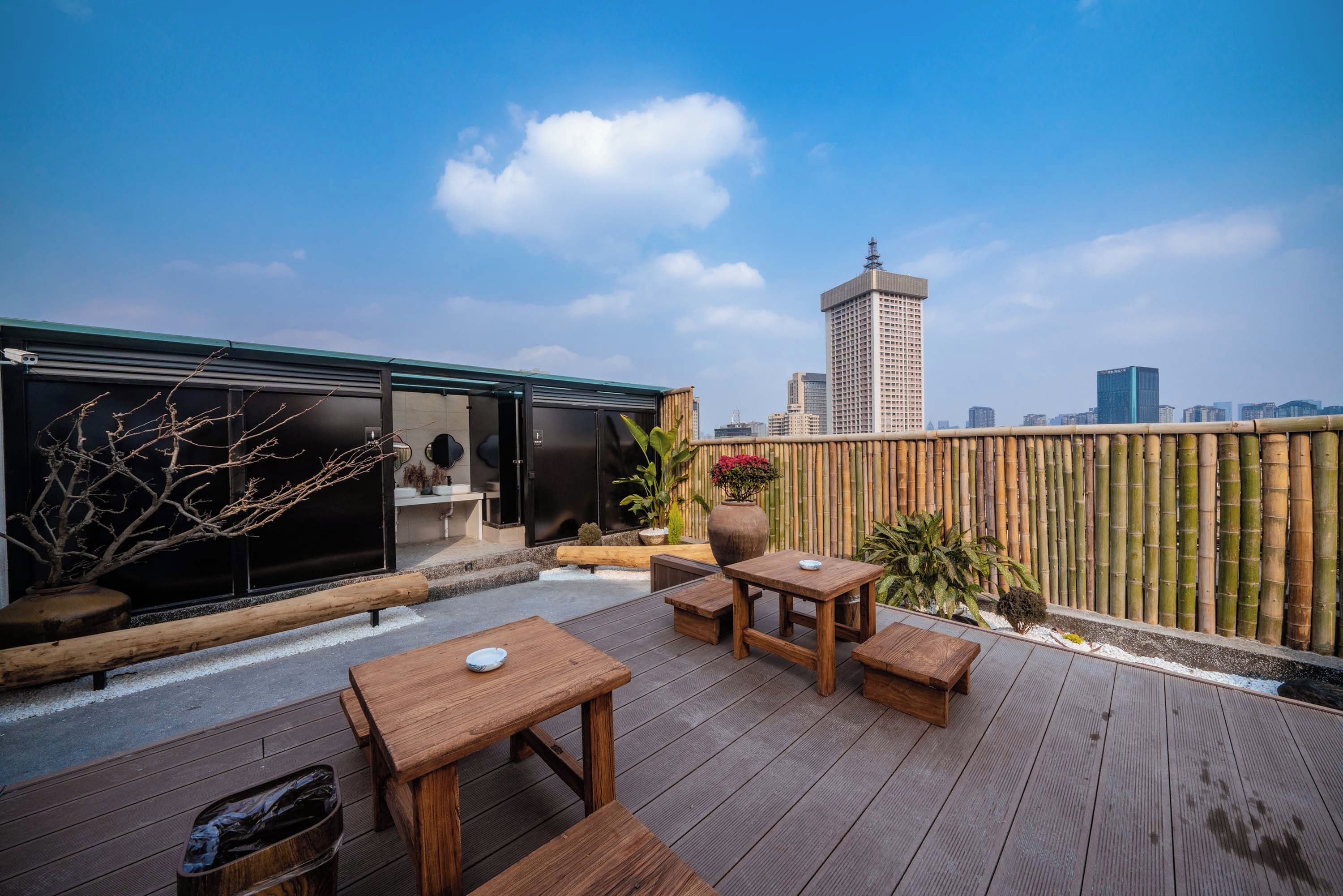 A rooftop terrace with wooden furniture, a view of the city skyline, and a bamboo fence.