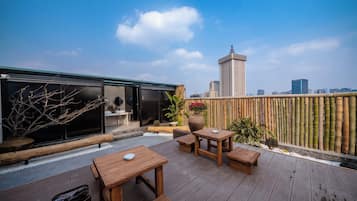 A rooftop terrace with wooden furniture, a view of the city skyline, and a bamboo fence.