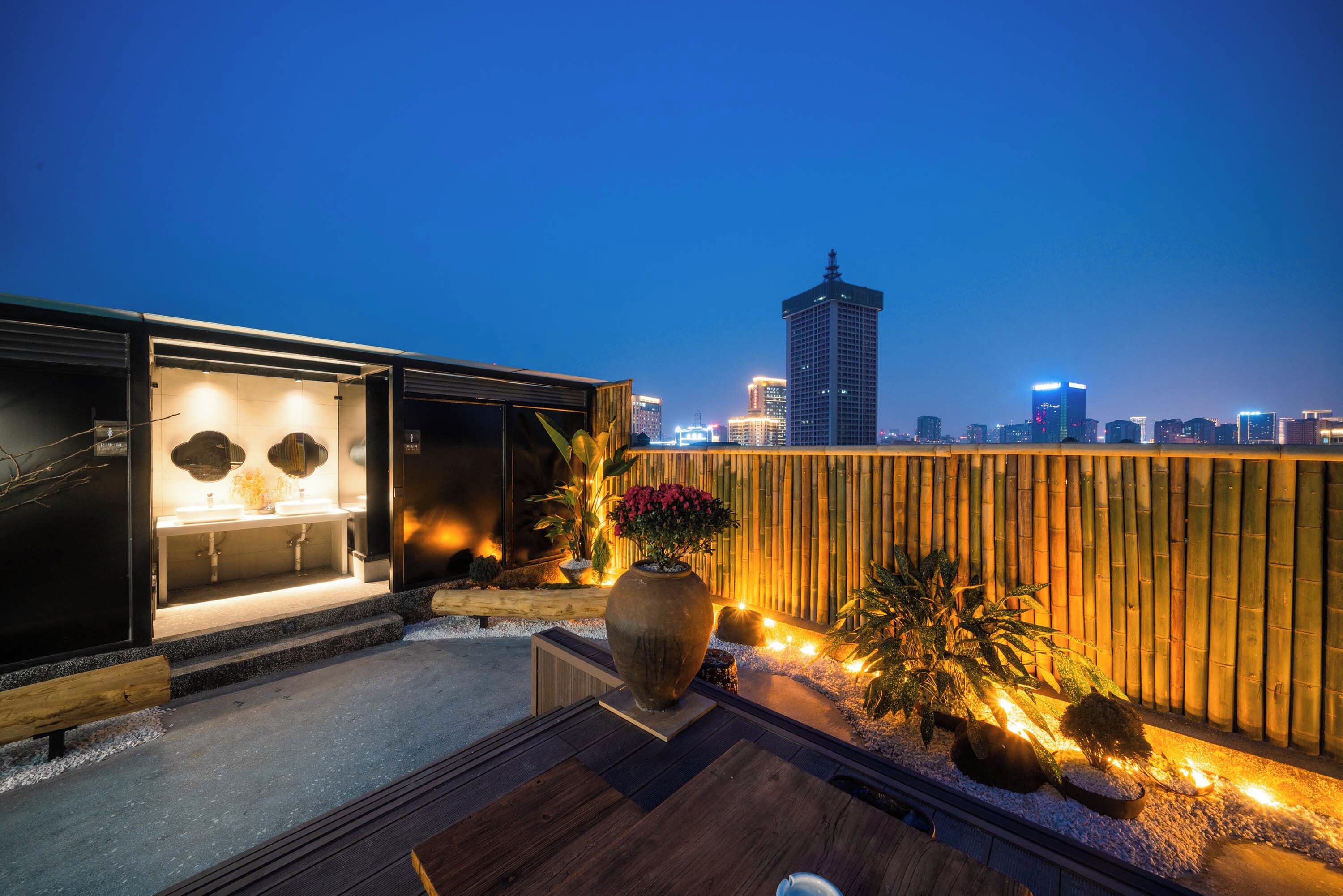 Una terraza en la azotea de diseño moderno, con zona de estar, una maceta grande con flores y vistas al horizonte de la ciudad al anochecer.