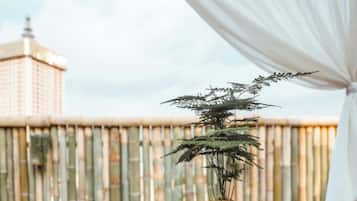 A bonsai tree on a wooden table with a view of a bamboo fence and a building in the background.
