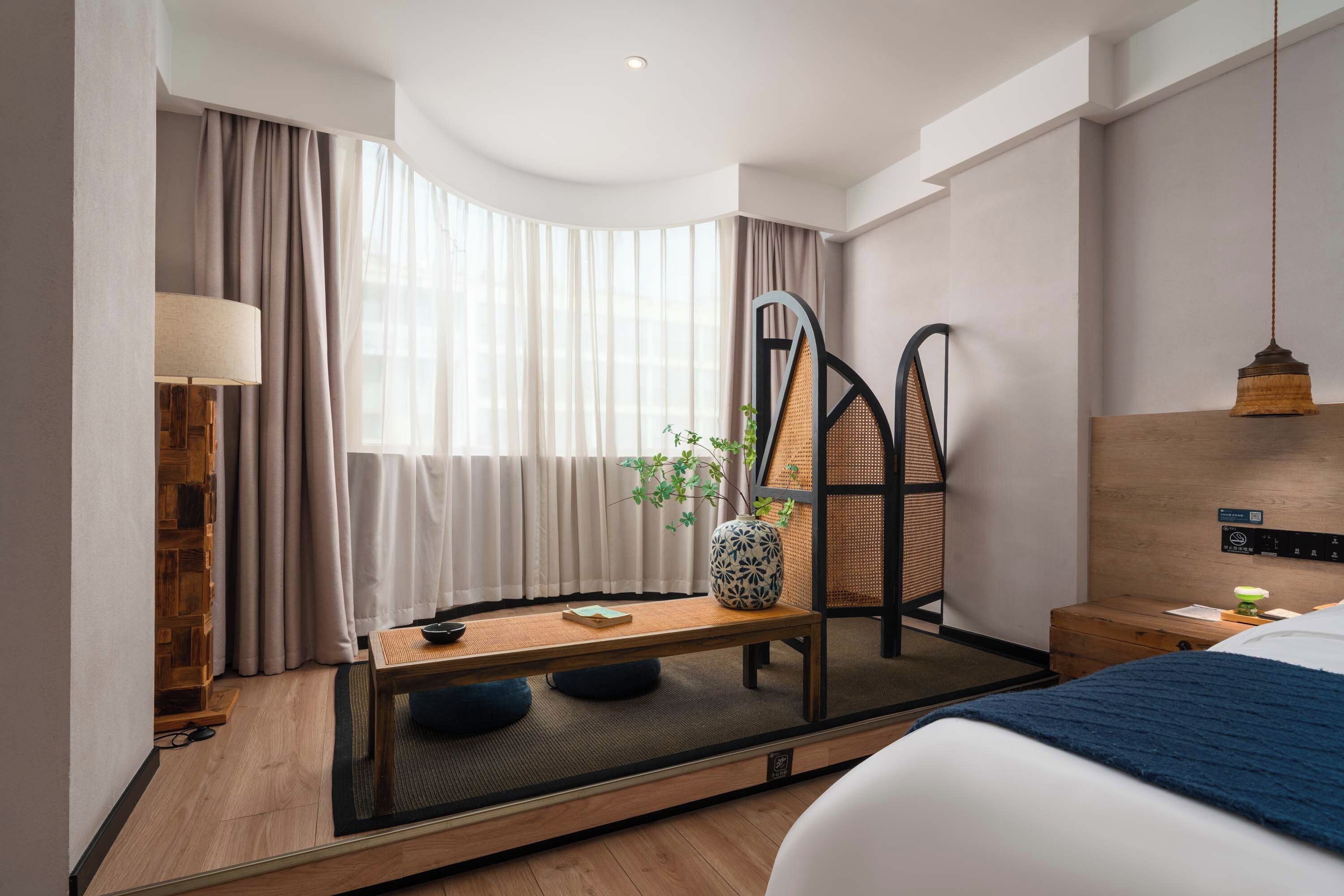 A modern hotel room with a bed, a desk, a chair, a lamp, and a window with curtains.