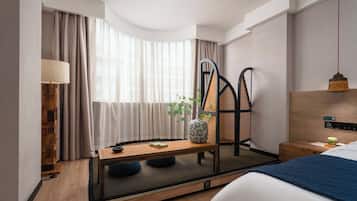 A modern hotel room with a bed, a desk, a chair, a lamp, and a window with curtains.