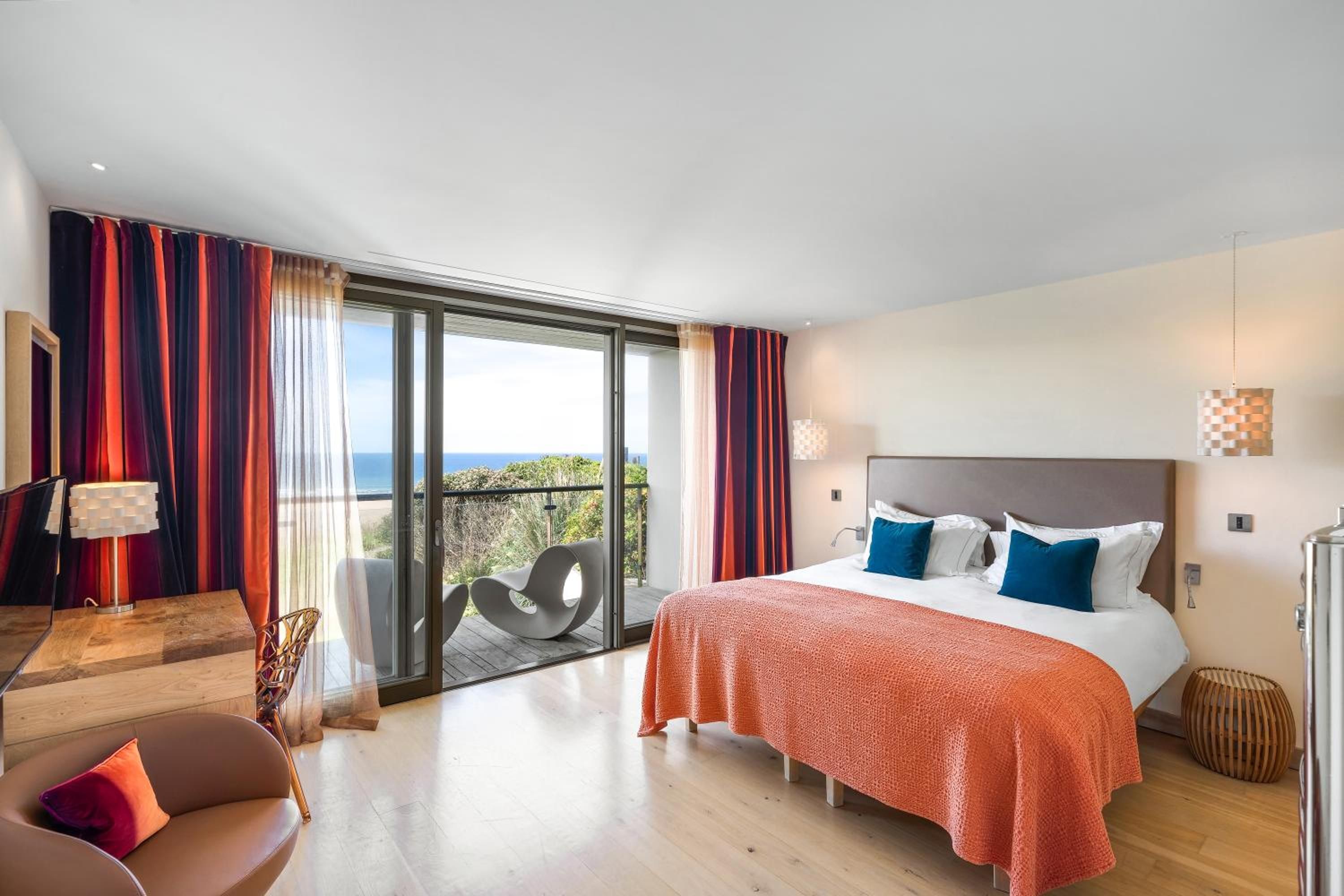 A modern hotel room with a large bed, a balcony with a view, and a seating area with a table and chairs.