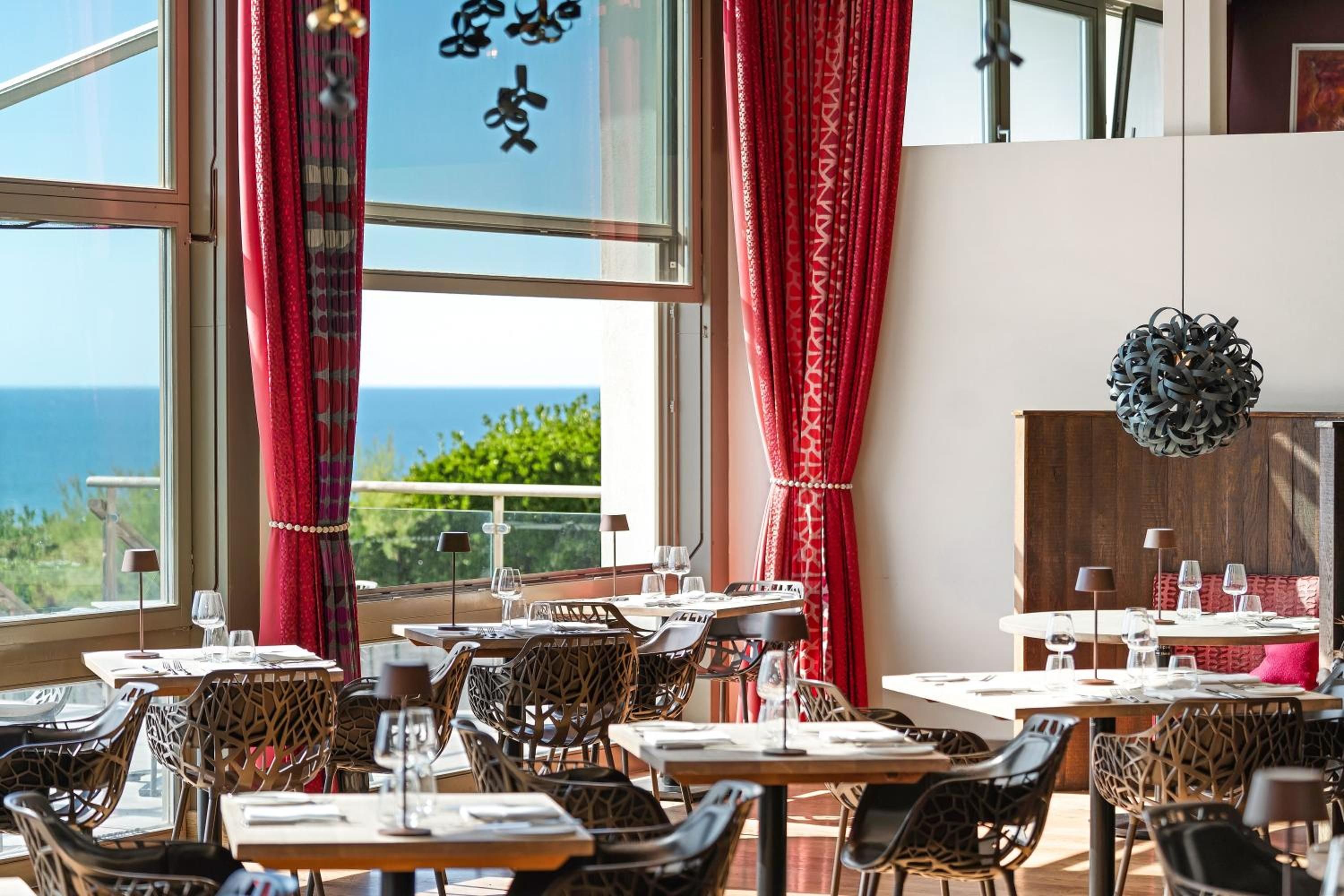 A restaurant with tables and chairs, large windows with red curtains, and a view of the sea.