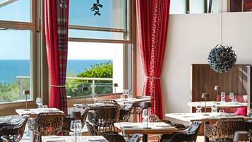 A restaurant with tables and chairs, large windows with red curtains, and a view of the sea.