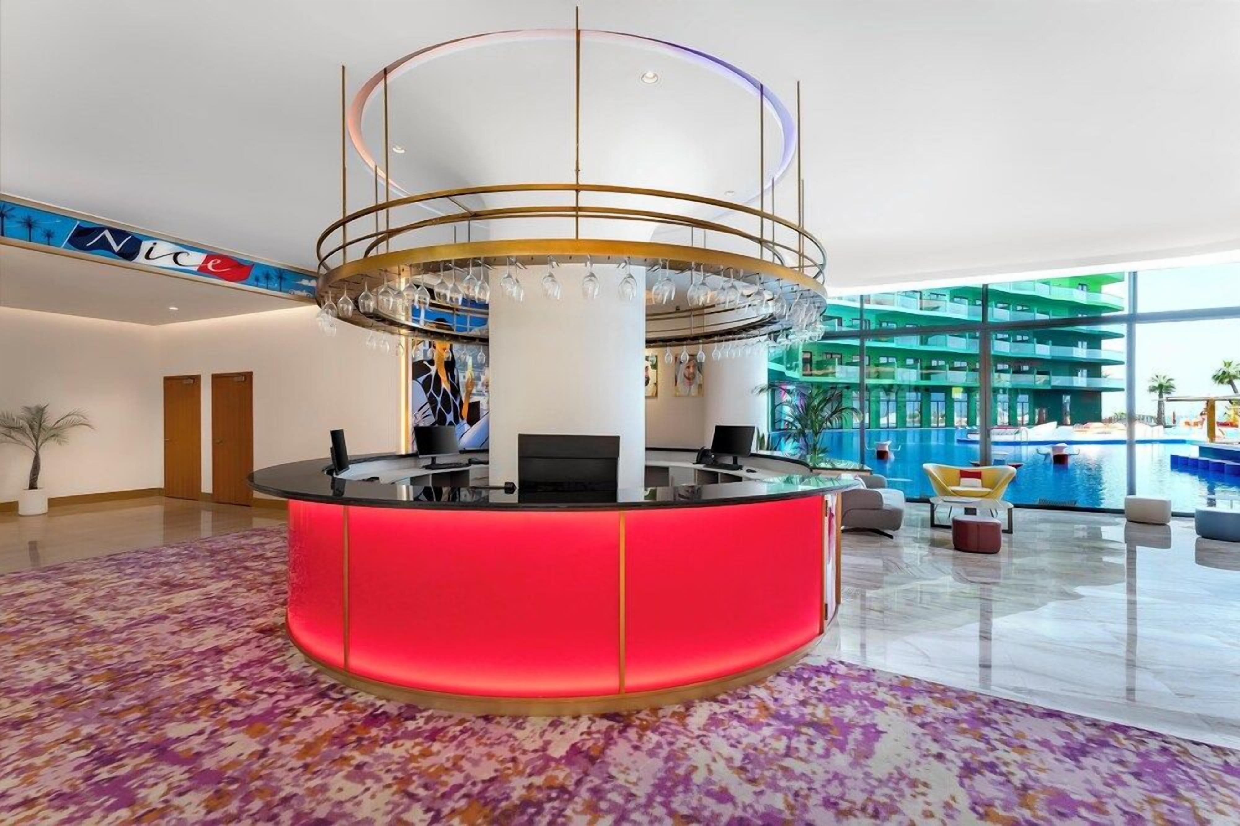 A hotel lobby with a central reception desk, a large chandelier, and a pool area visible through the glass walls.