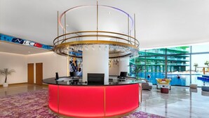 Een hotellobby met een centraal receptiebalie, een grote kroonluchter en een zwembadgedeelte zichtbaar door glazen wanden.