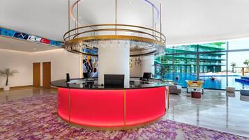 A hotel lobby with a central reception desk, a large chandelier, and a pool area visible through the glass walls.