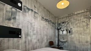 Hotellihuone, jossa on suuri sänky, yöpöytä, tuoli, seinään kiinnitetty valaisin ja sisäänrakennettu hylly.