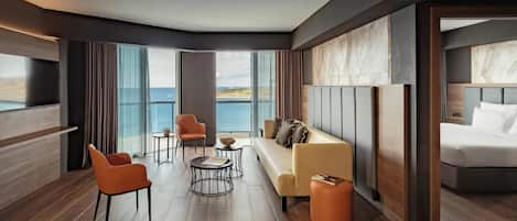 A modern hotel room with a large bed, a sofa, two armchairs, a small round table, and a view of the sea.