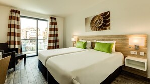 Habitación de hotel con una cama grande, dos mesitas de noche, un velador, una ventana con cortinas y vistas a la ciudad, además de una obra de arte colgada en la pared.