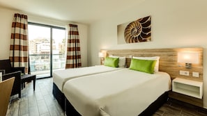 Habitació d'hotel amb un llit gran, dues taules de nit, una tauleta auxiliar, una finestra amb cortines, vista a la ciutat i una obra d'art penjada a la paret.