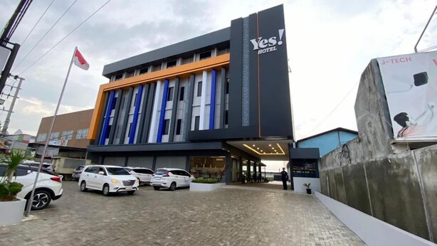 A modern hotel with a prominent entrance, a parking area, and a flag. - Yes Hotel (Bandar Lampung)