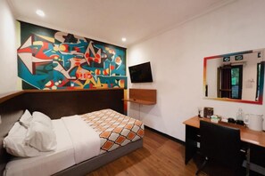 Habitación de hotel con cama, escritorio, silla, espejo y televisión montada en la pared.