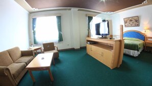 Ein Zimmer mit einer Couch, einem Couchtisch, einem Fernseher auf einem Schrank, einem Bett mit blauer Kopfteil und einem Fenster mit Vorhängen.