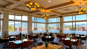 A dining area with multiple tables and chairs, large windows with a view of a cityscape and mountains, and decorative chandeliers. - Atiyan Hotel Syariah (Bukittinggi)