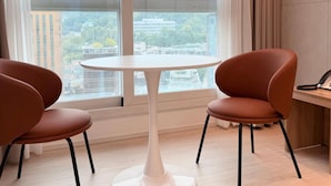 Ein modernes Zimmer mit einem runden Tisch und zwei Stühlen, großen Fenstern mit Blick auf die Stadt und einem Vorhang auf der rechten Seite.