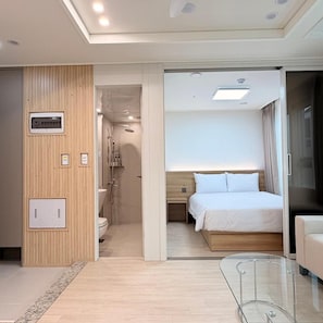 Habitació d'hotel moderna amb una taula de cafè de vidre, un llit amb roba de llit blanca, un panell de capçal de fusta i un bany amb dutxa i vàter.