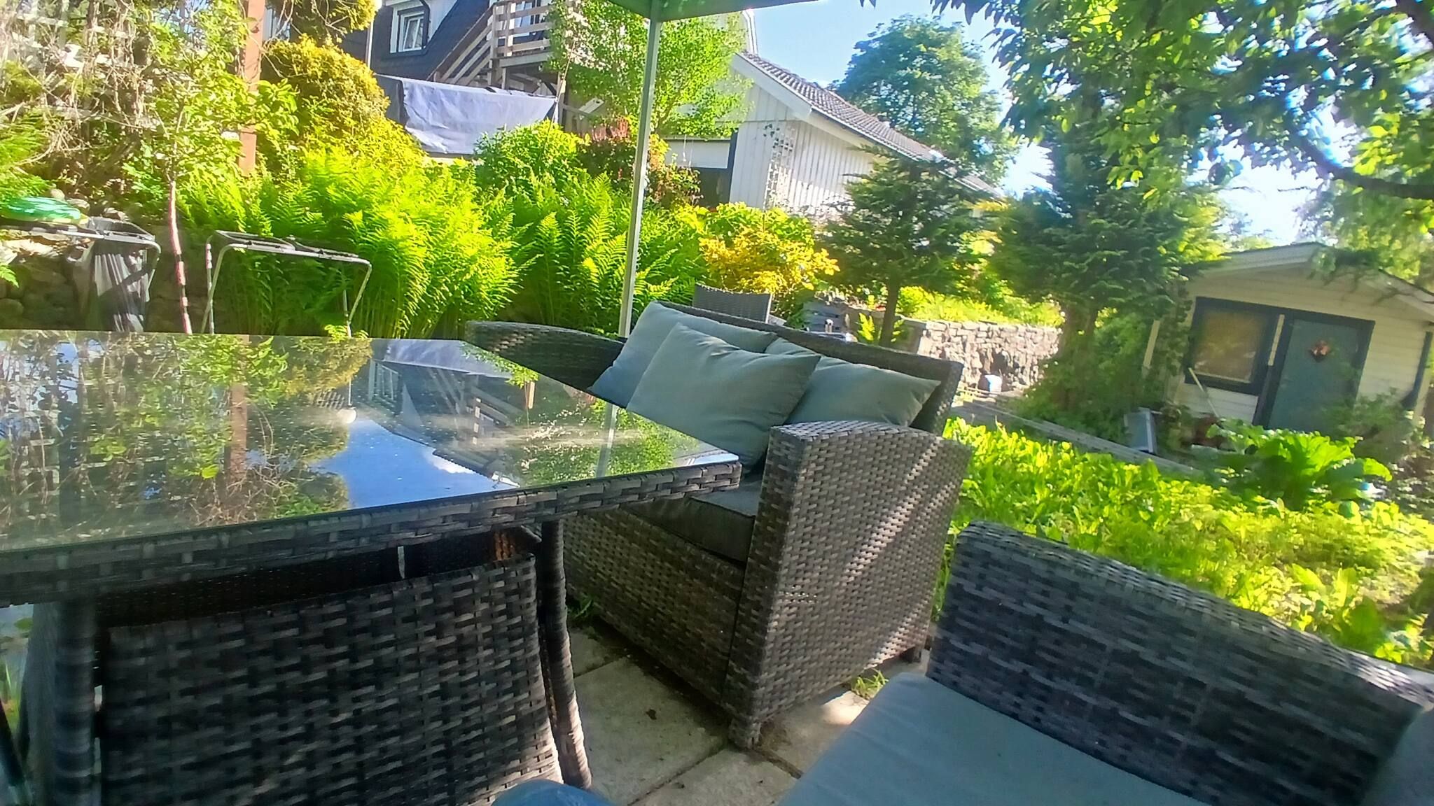 Un patio al aire libre con mobiliario de mimbre, una mesa de vidrio y vistas a un jardín y edificios al fondo.