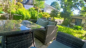 An outdoor patio with wicker furniture, a glass table, and a view of a garden and buildings in the background.