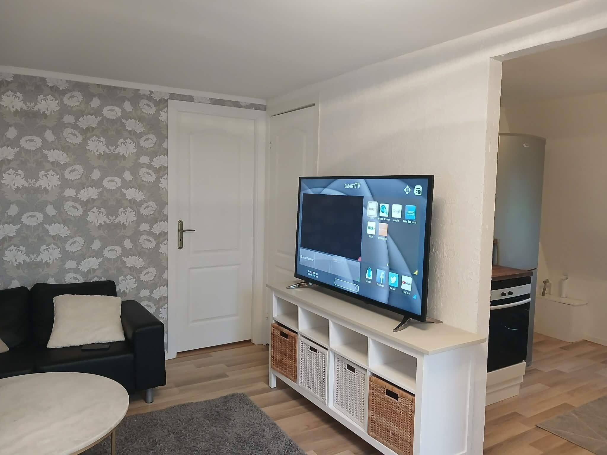 En stue med et fladskærms-tv på et stativ, en sofa, et sofabord og en dør med blomstret tapet.